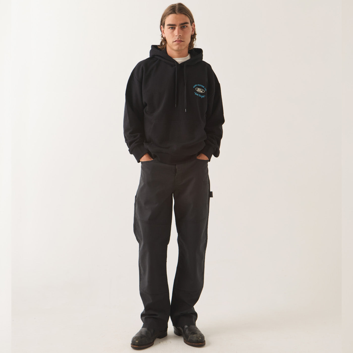 A model stands against a plain light background wearing a black pullover hoodie with a small blue and white graphic on the left chest, paired with dark charcoal cargo pants and black shoes.