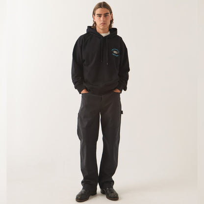A model stands against a plain light background wearing a black pullover hoodie with a small blue and white graphic on the left chest, paired with dark charcoal cargo pants and black shoes.