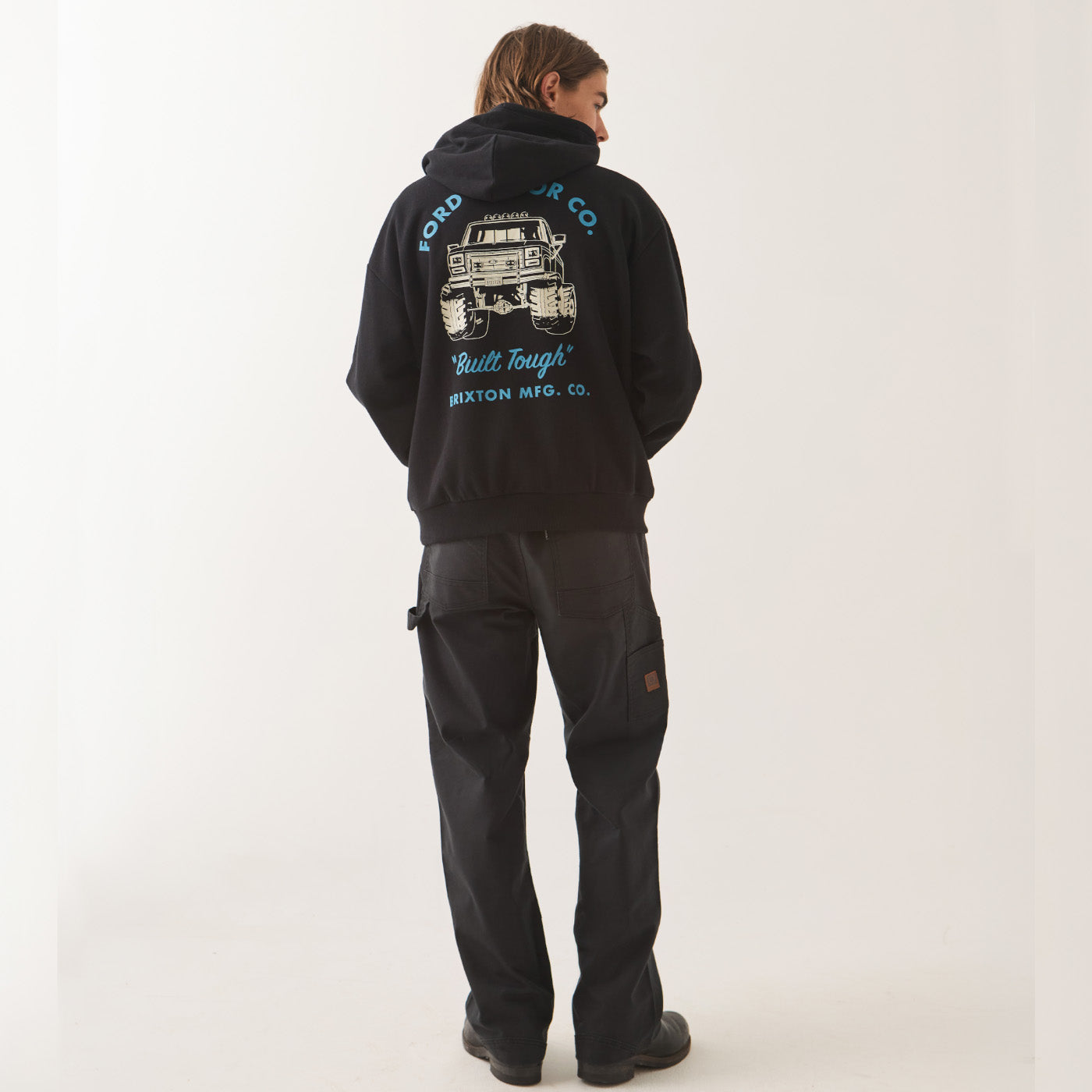 A model viewed from behind wearing a black Ford Ranger hoodie with a large graphic print on the back, paired with black cargo pants, against a plain off-white studio background.