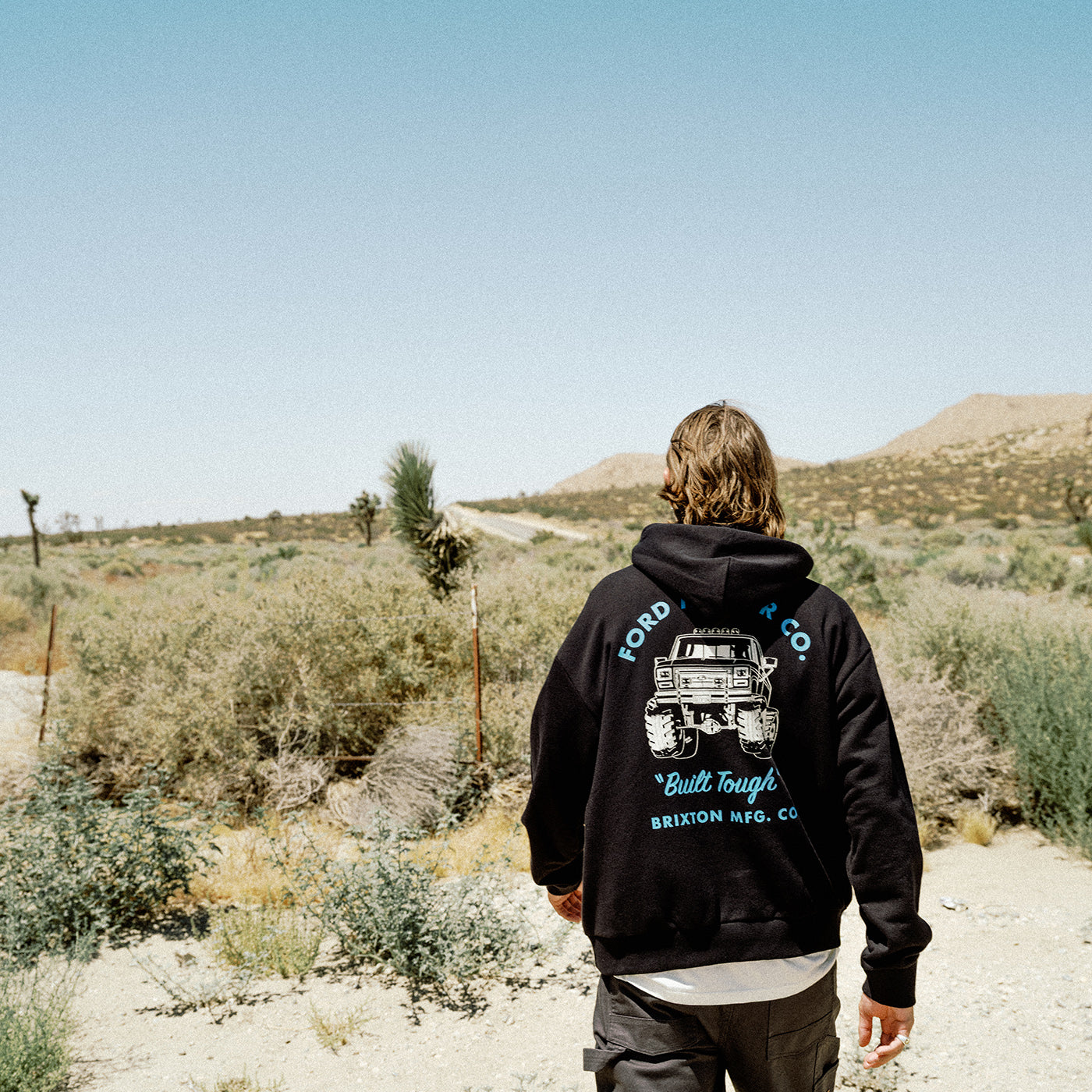 A person viewed from behind wearing a black hoodie with a large back graphic of a vintage off-road vehicle and the text 'FORD 500 CO' and 'Built Tough Brixton MD, CO', standing in a desert landscape.