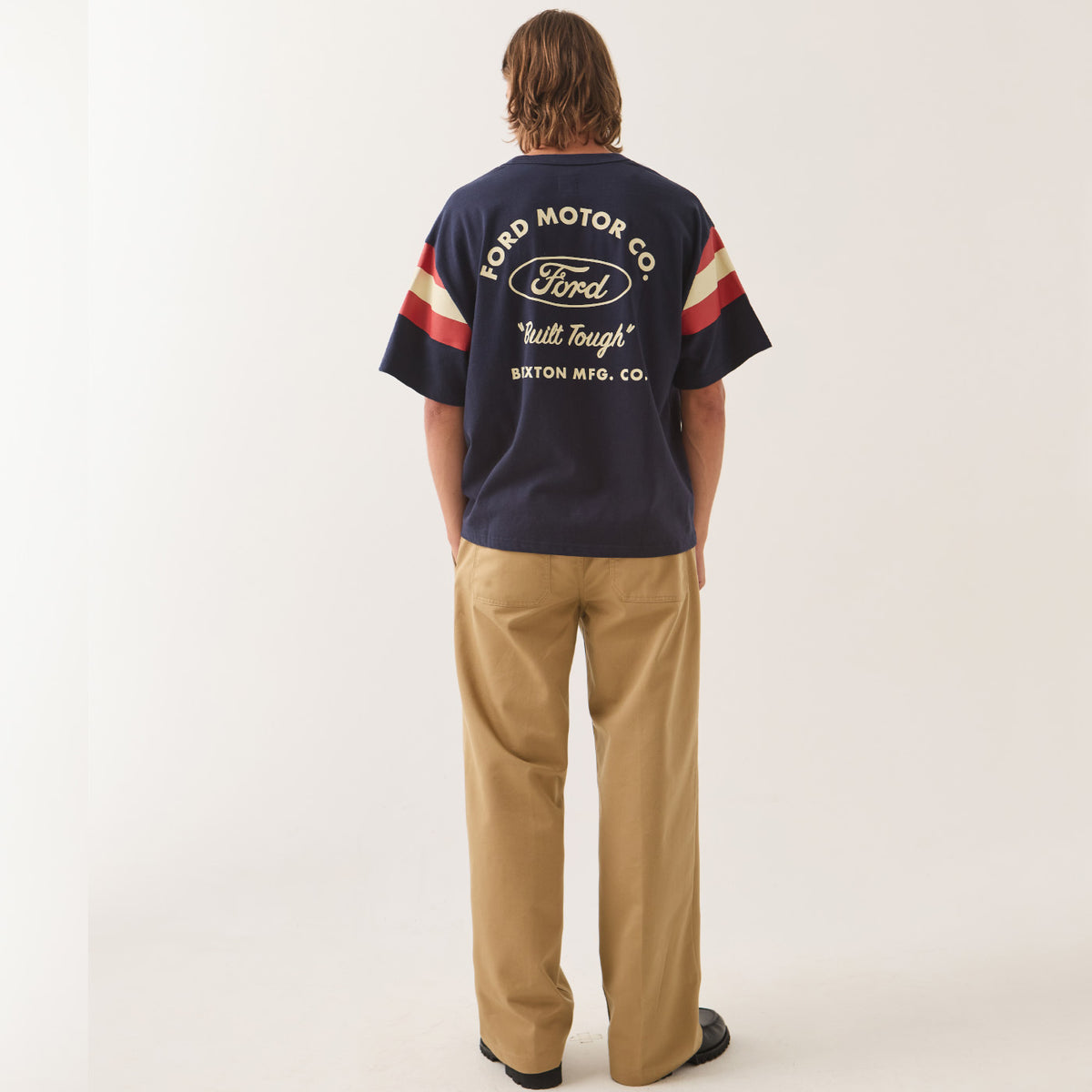 A man viewed from behind wearing a navy blue Ford Motor Co. t-shirt with cream and red sleeve stripes, paired with tan trousers, against a plain off-white studio background.
