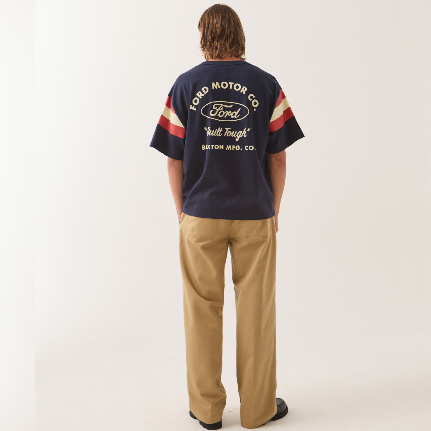 A man viewed from behind wearing a navy blue Ford Motor Co. t-shirt with cream and red sleeve stripes, paired with tan trousers, against a plain off-white studio background.