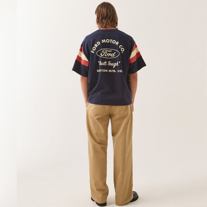 A man viewed from behind wearing a navy blue Ford Motor Co. t-shirt with cream and red sleeve stripes, paired with tan trousers, against a plain off-white studio background.