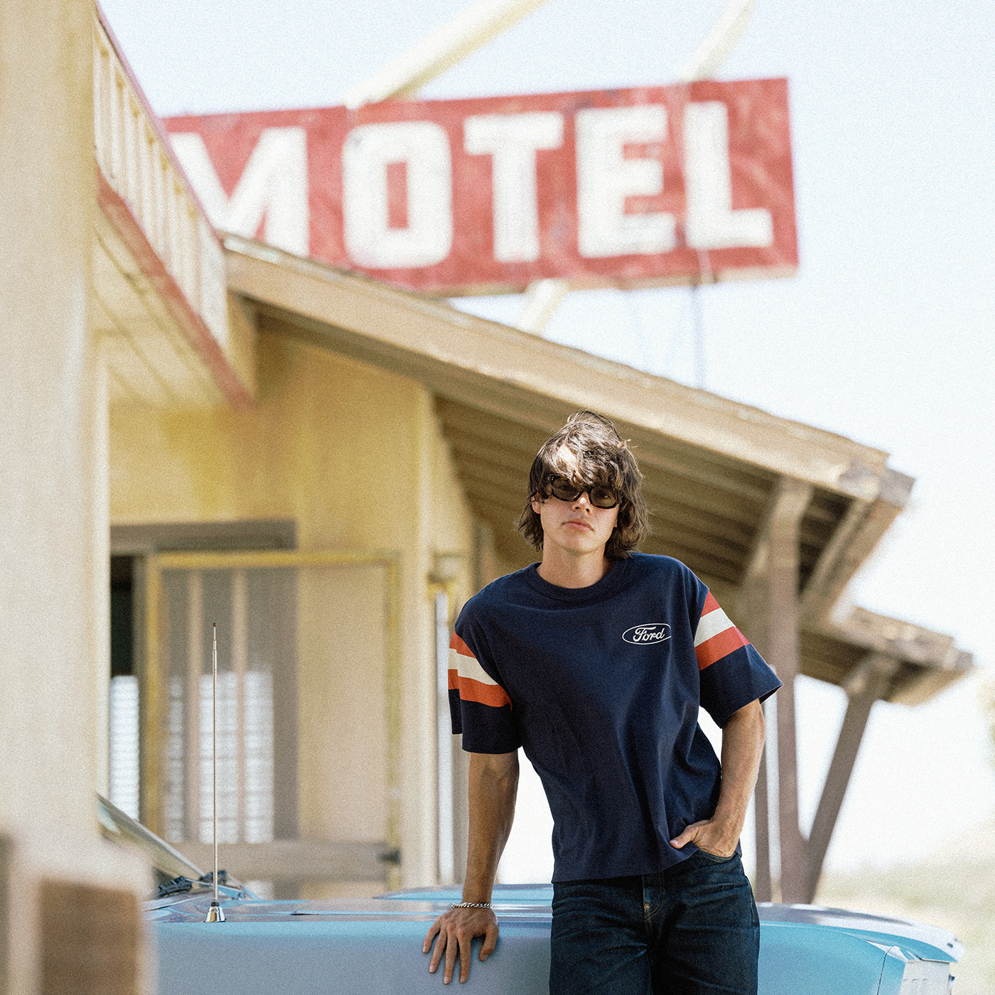 A man wearing a navy blue Ford t-shirt with orange and white sleeve stripes leans against a light blue car in front of a motel sign.