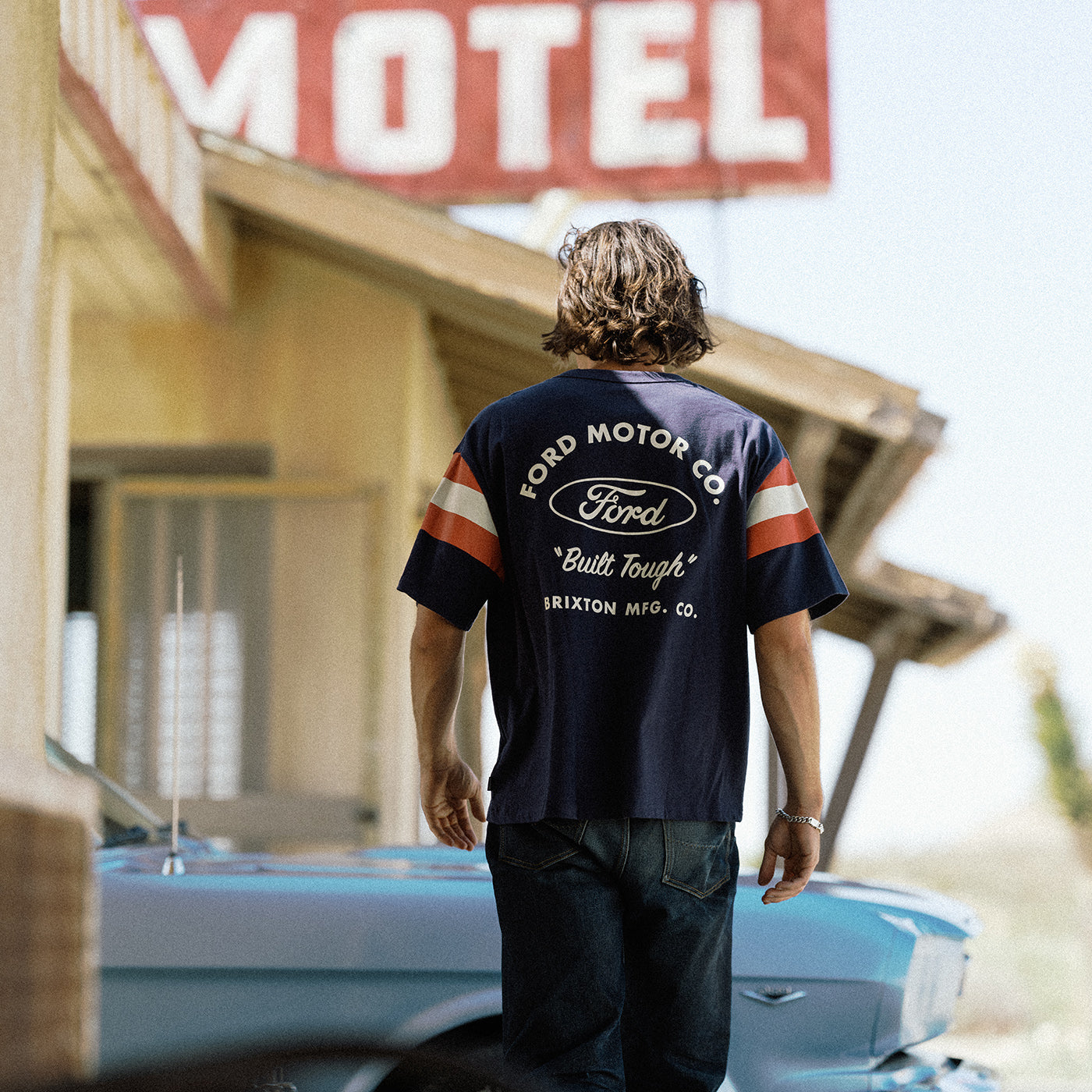 A man viewed from behind wearing a navy blue Ford Motor Co. t-shirt with orange and white sleeve stripes, standing in front of a vintage motel sign and a light blue car.