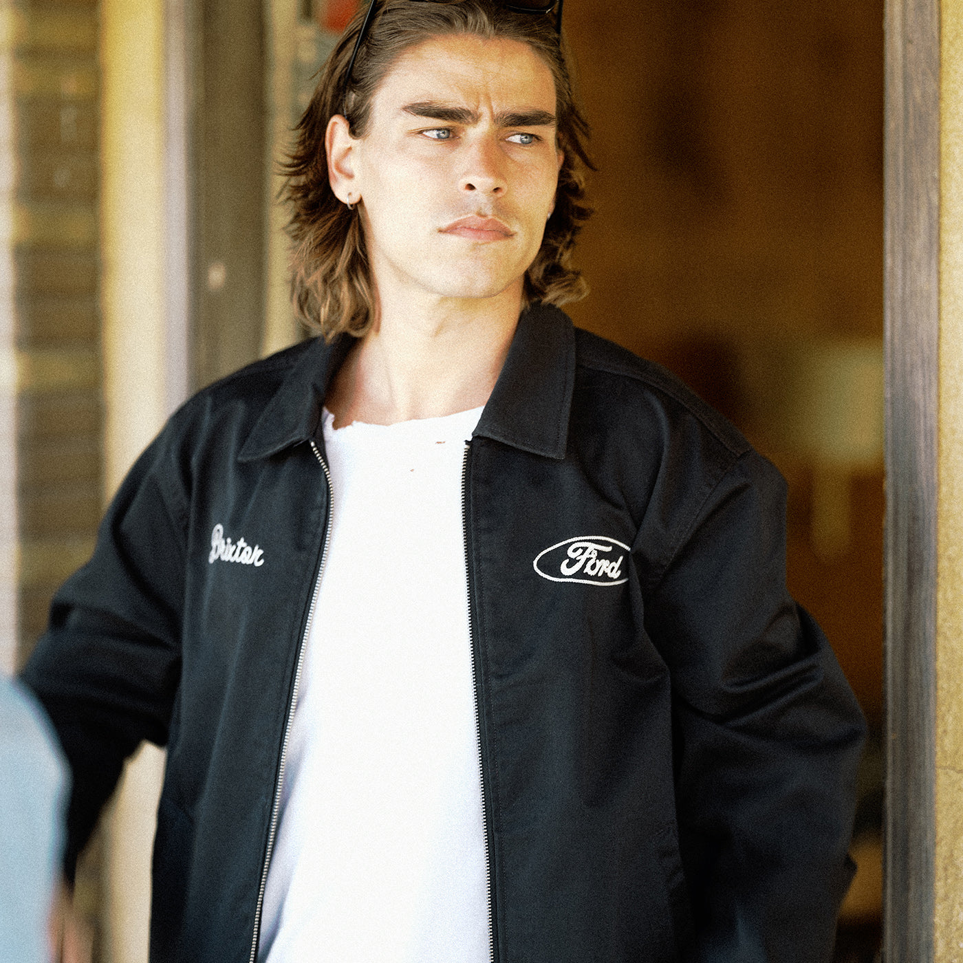 A man wearing a black zip-up jacket with 'Bastak' embroidered on the right chest and a Ford logo on the left chest, over a white t-shirt.