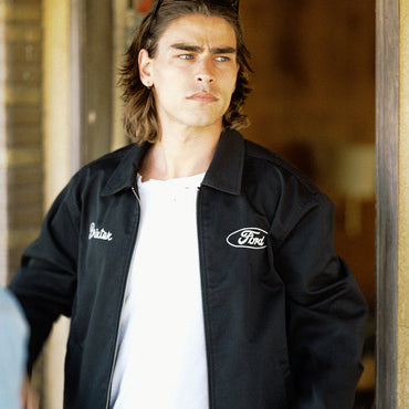 A man wearing a black zip-up jacket with 'Bastak' embroidered on the right chest and a Ford logo on the left chest, over a white t-shirt.