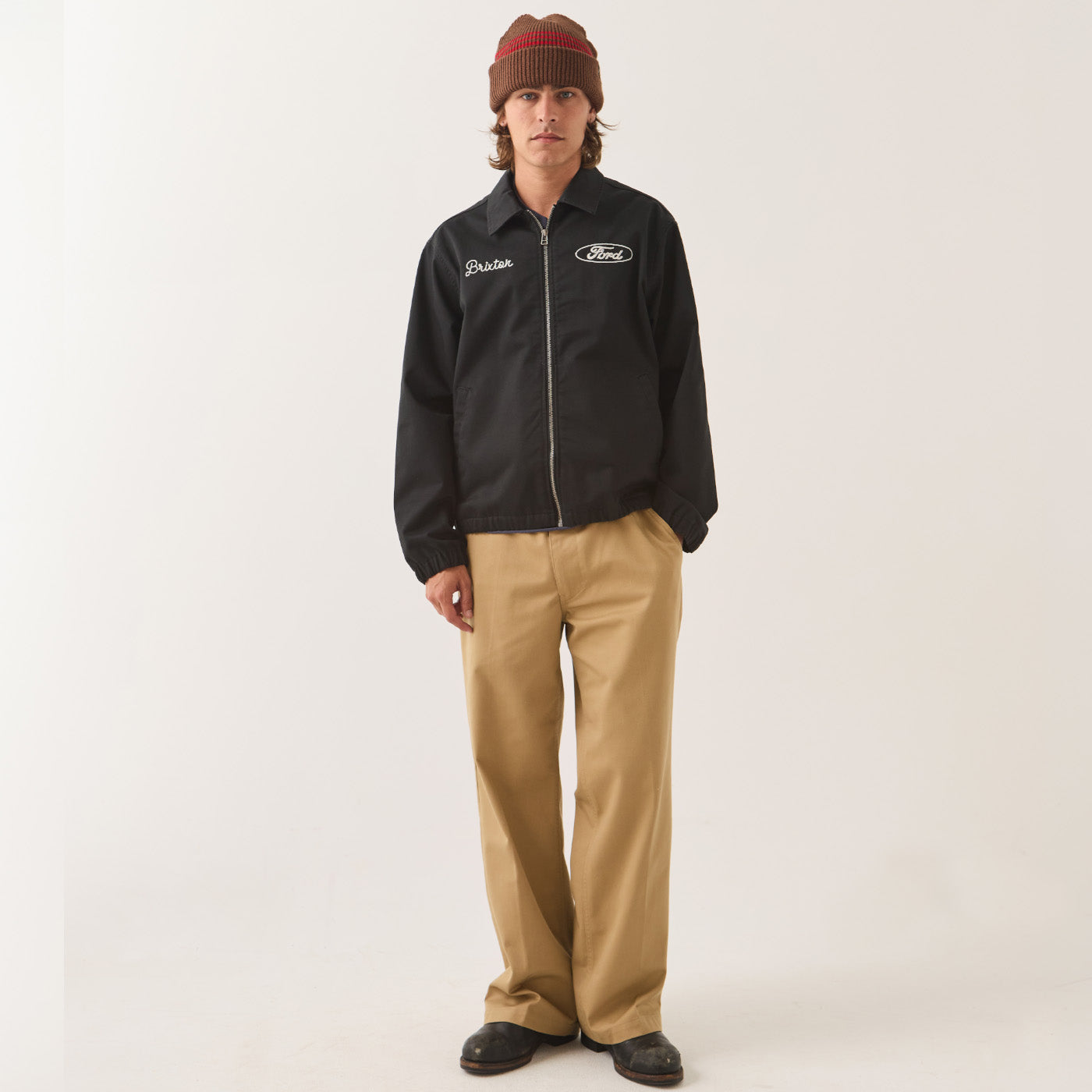 A model stands against a plain light background wearing a black Ford zip-up jacket with white embroidered logos, tan pants, and a brown knit beanie.