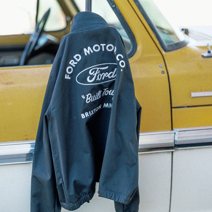 A black Ford jacket with white embroidered lettering on the back, hanging on a post in front of a vintage yellow and white car.