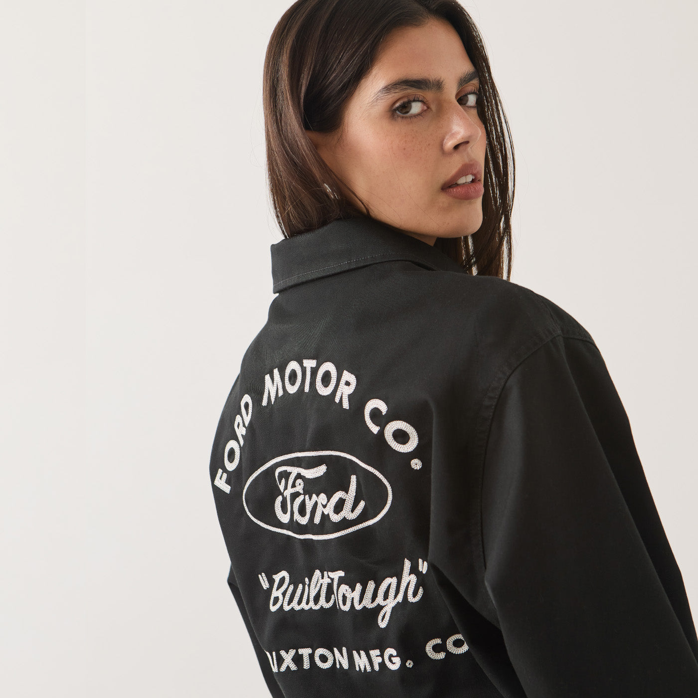 A woman wearing a black Ford jacket, shown from the back over her shoulder. The jacket features a large white embroidered logo and text on the back, including 'FORD MOTOR CO.