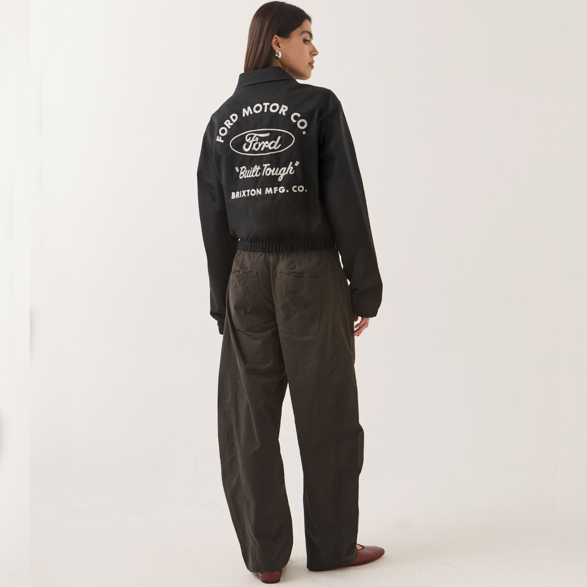 A woman shown from the back wearing a black cropped jacket with a large white Ford Motor Co. logo and text on the back, paired with dark wide-leg pants and brown shoes, against a plain light background.