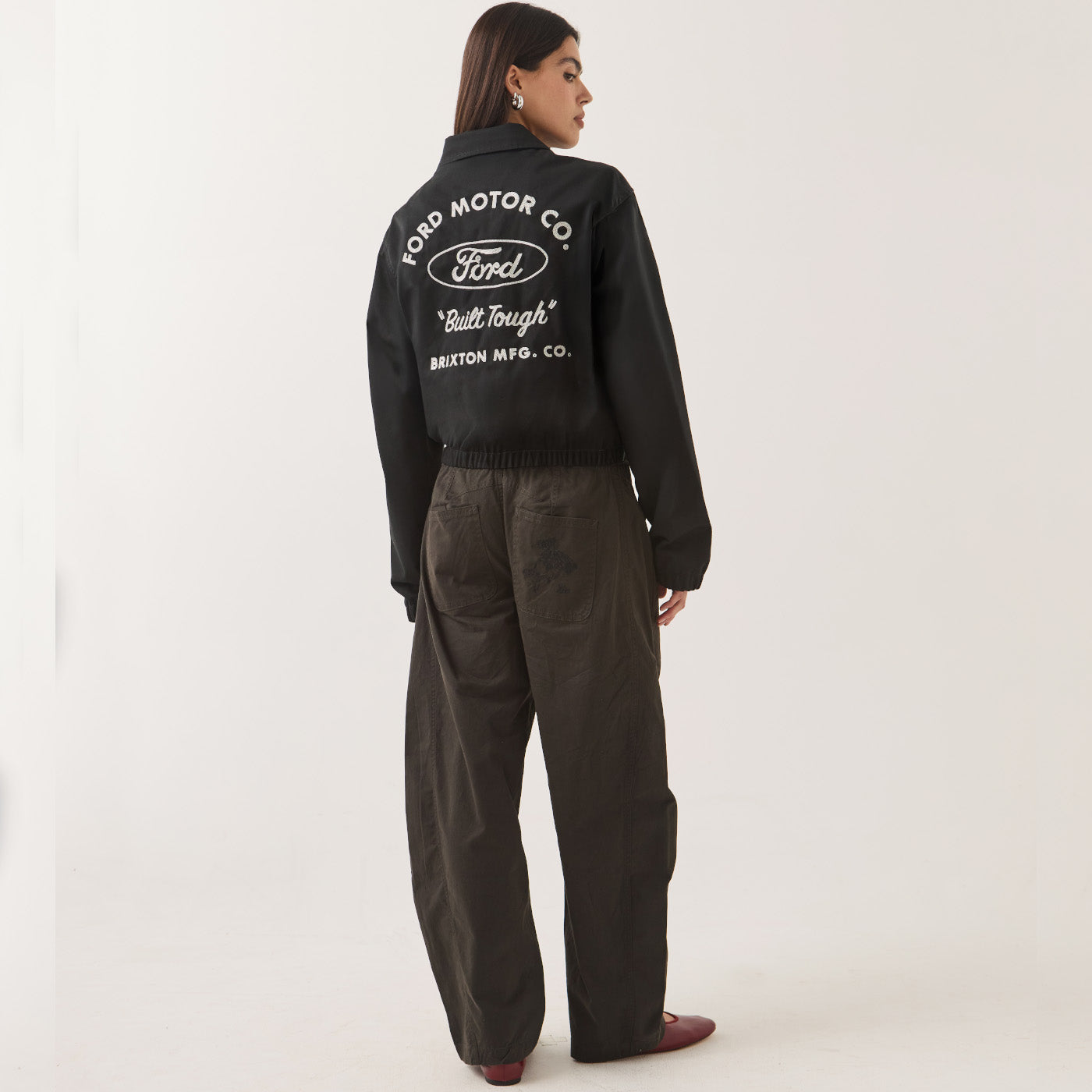 A woman shown from the back wearing a black cropped jacket with a large white Ford Motor Co. logo and text on the back, paired with dark wide-leg pants and brown shoes, against a plain light background.