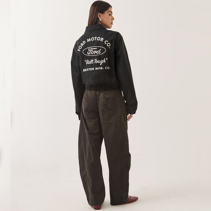 A woman shown from the back wearing a black cropped jacket with a large white Ford Motor Co. logo and text on the back, paired with dark wide-leg pants and brown shoes, against a plain light background.