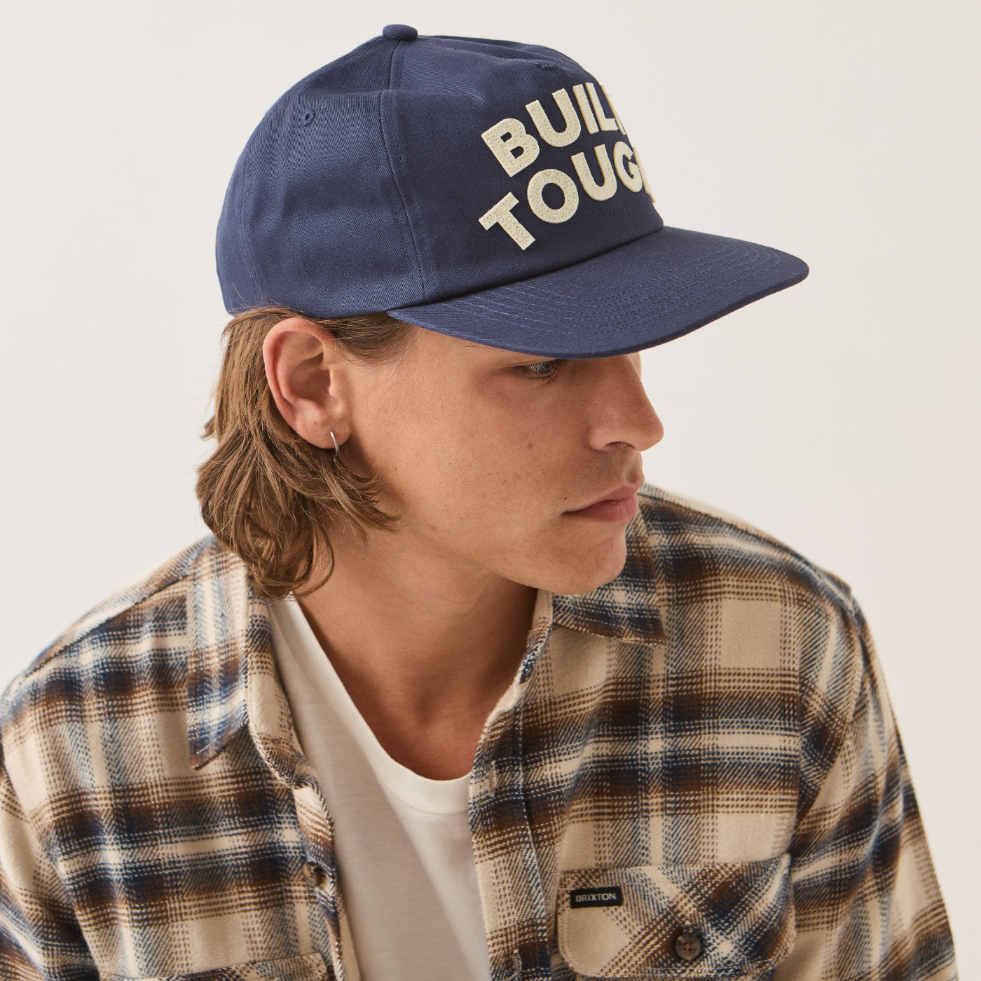 A man in profile wearing a navy blue baseball cap with 'BUILD TOUGH' embroidered in cream on the front, paired with a beige and navy plaid flannel shirt over a white t-shirt, against a plain light background.