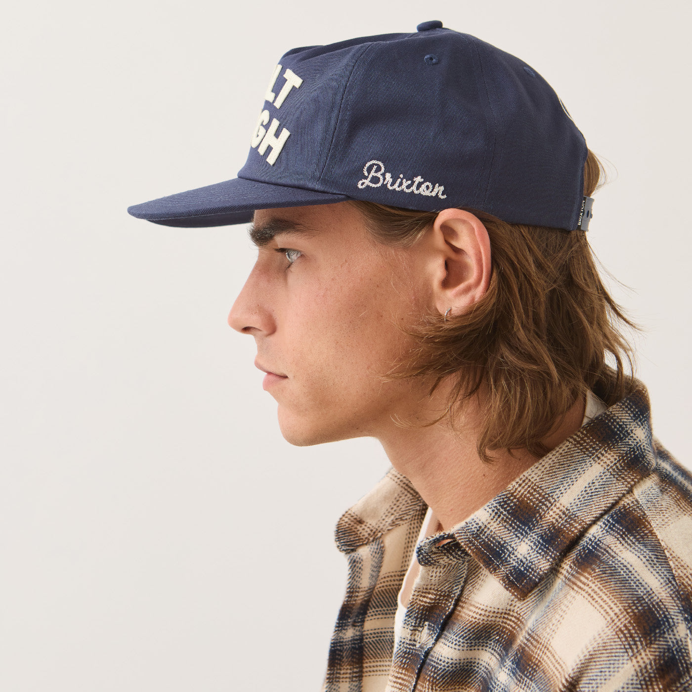 A man in profile wearing a navy blue Brixton baseball cap with white embroidered lettering, paired with a brown and beige plaid flannel shirt, against a light gray background.