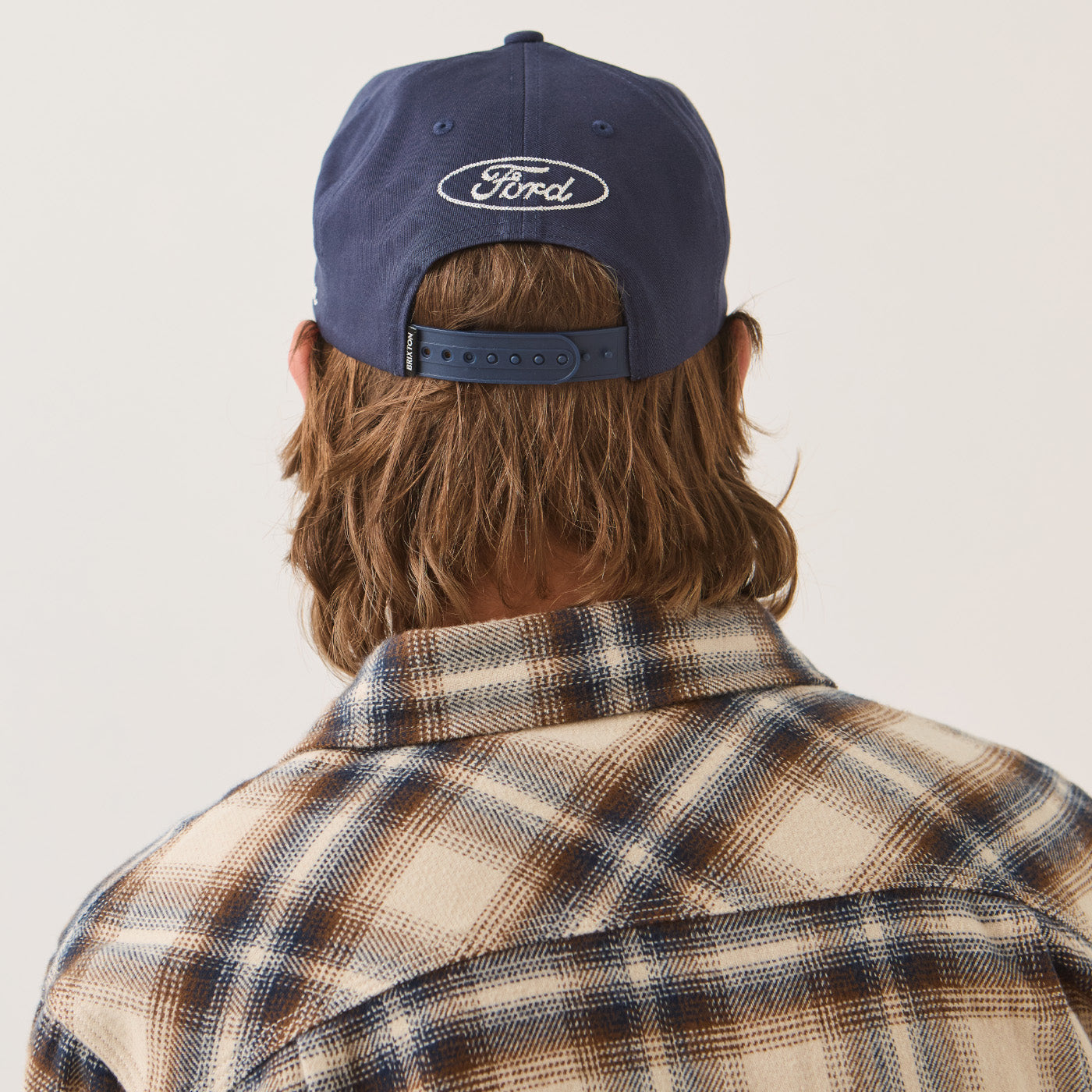 Rear view of a person wearing a navy blue Ford baseball cap and a brown and beige plaid flannel shirt against a light gray background.
