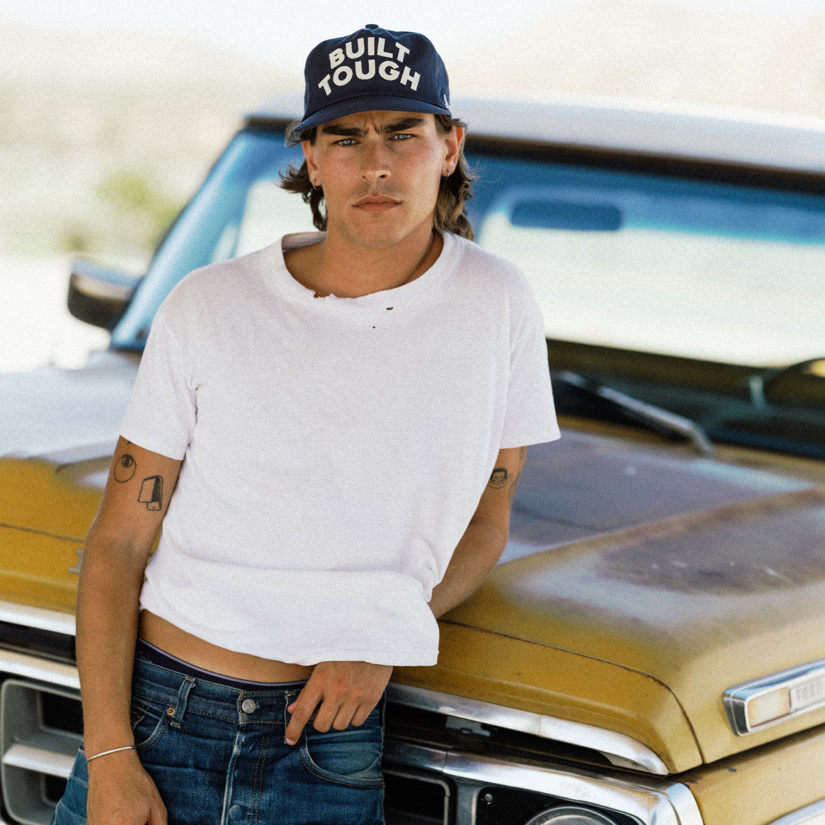 A man wearing a white t-shirt, blue jeans, and a navy blue cap with 'BUILT TOUGH' in white text leans against the front of a vintage gold truck.