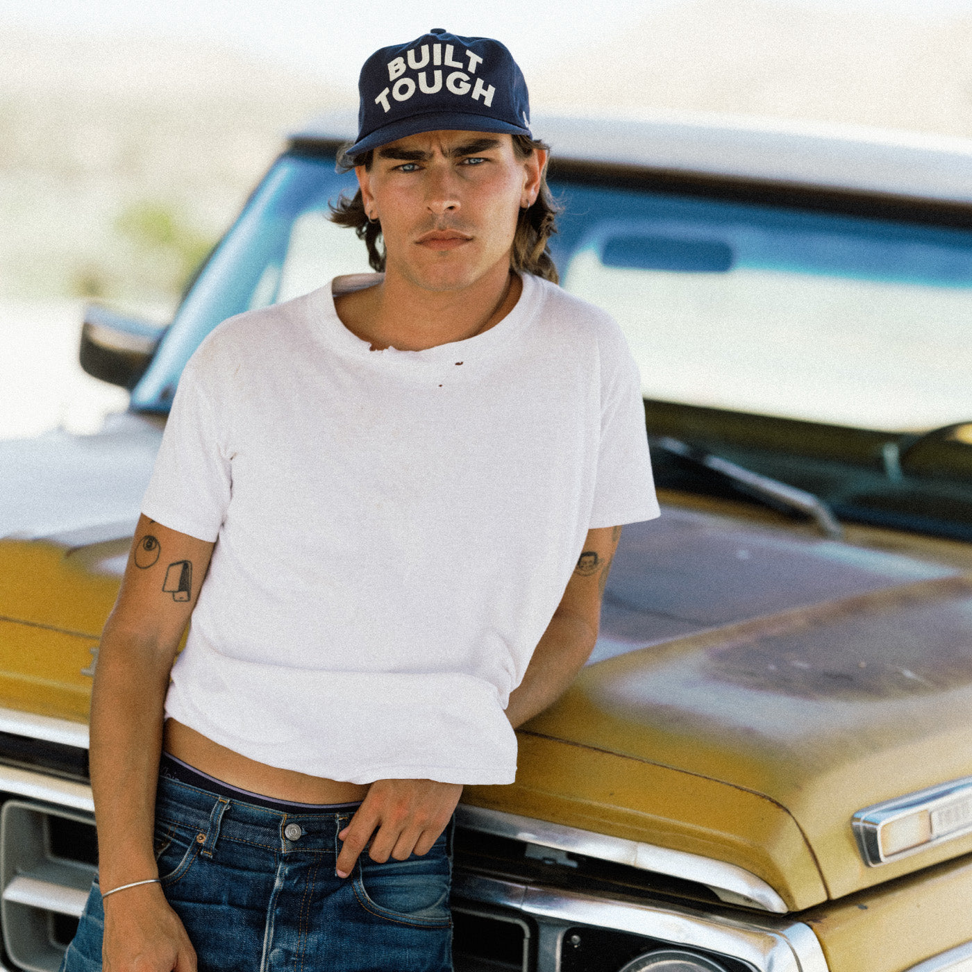 A man wearing a white t-shirt, blue jeans, and a navy blue cap with 'BUILT TOUGH' in white text leans against the front of a vintage gold truck.