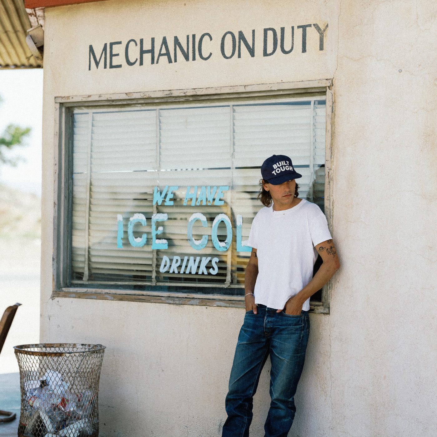 A man wearing a white t-shirt, blue jeans, and a dark baseball cap with 'MECHANIC ON DUTY' stands leaning against the exterior wall of a building with a window displaying 'ICE COOL DRINKS'.