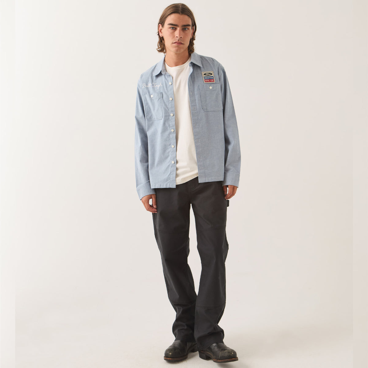 A man modeling a light blue denim button-up shirt with a colorful patch on the left chest, worn over a white t-shirt and dark gray pants, against a plain light gray studio background.