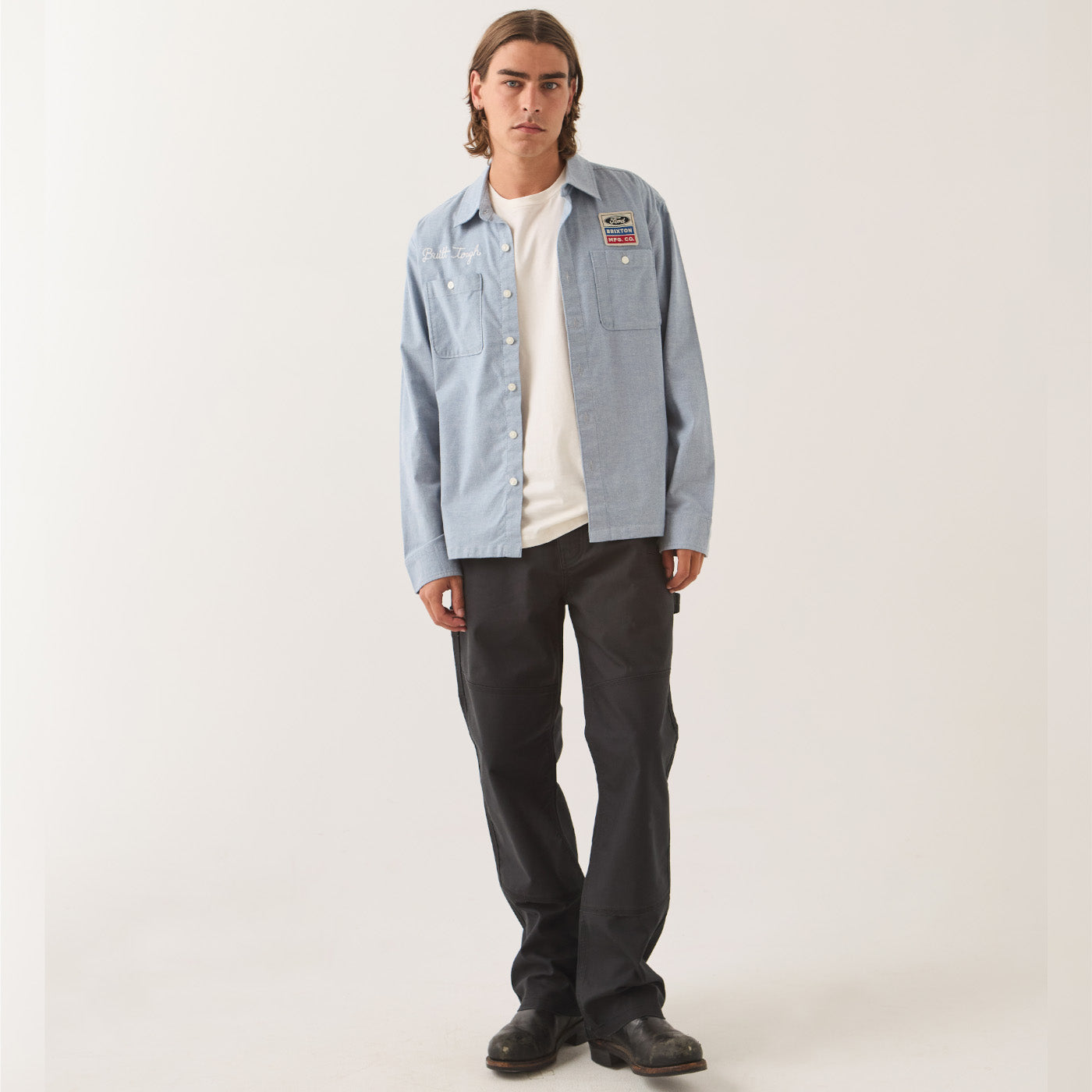 A man modeling a light blue denim button-up shirt with a colorful patch on the left chest, worn over a white t-shirt and dark gray pants, against a plain light gray studio background.