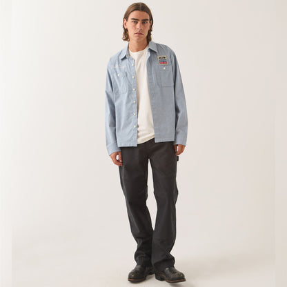 A man modeling a light blue denim button-up shirt with a colorful patch on the left chest, worn over a white t-shirt and dark gray pants, against a plain light gray studio background.
