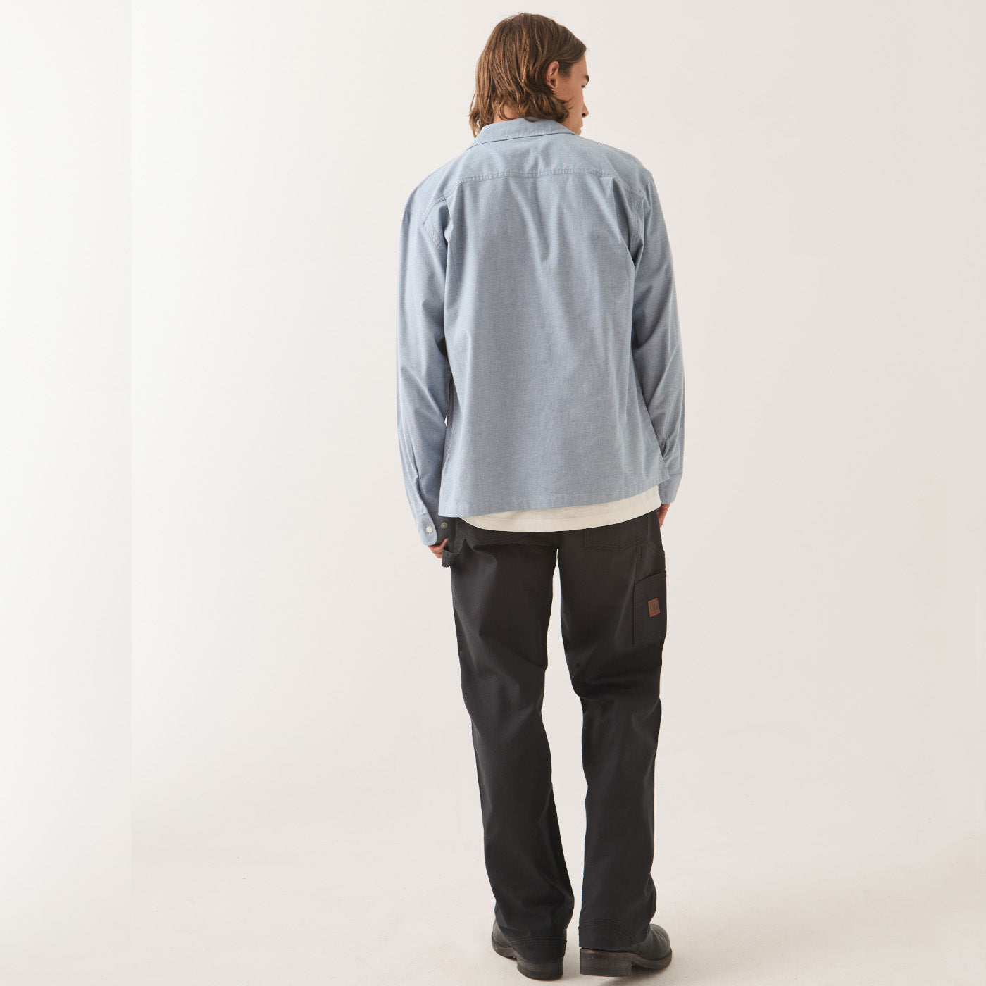 A model shown from the back wearing a light blue long-sleeve button-up shirt, black pants, and black shoes, standing against a plain off-white studio background.