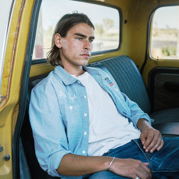A man sitting in the back of a yellow vintage truck wearing a light blue button-up shirt with white embroidery over a white t-shirt and blue jeans.