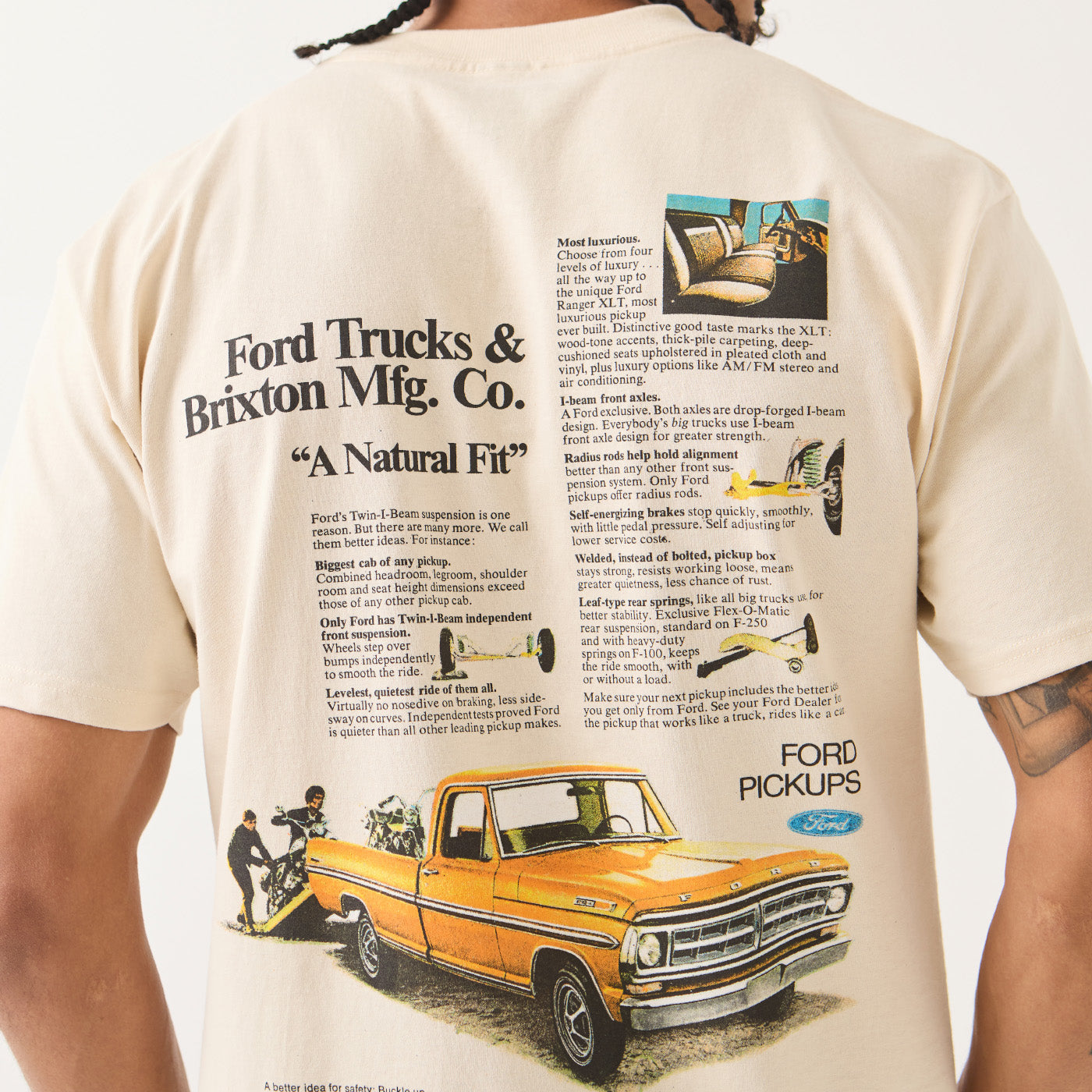 A person wearing a cream-colored graphic t-shirt featuring a vintage Ford Trucks & Brixton Mfg. Co.