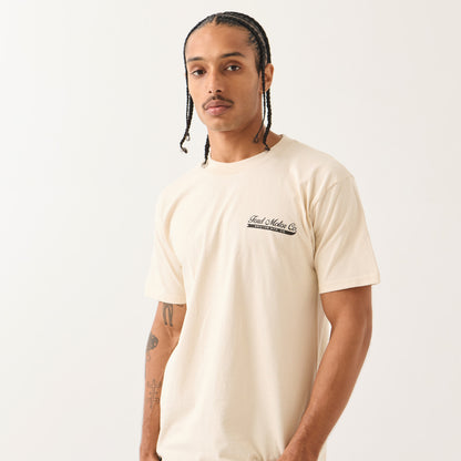 A man wearing a cream-colored short-sleeve t-shirt with a black 'Fuel Makers Co' logo printed on the left chest, photographed against a clean off-white background.
