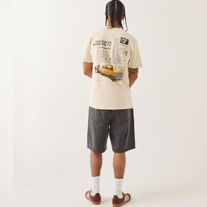 A man is shown from behind wearing a cream-colored graphic t-shirt, dark gray denim shorts, white socks, and brown sneakers against a plain white studio background.