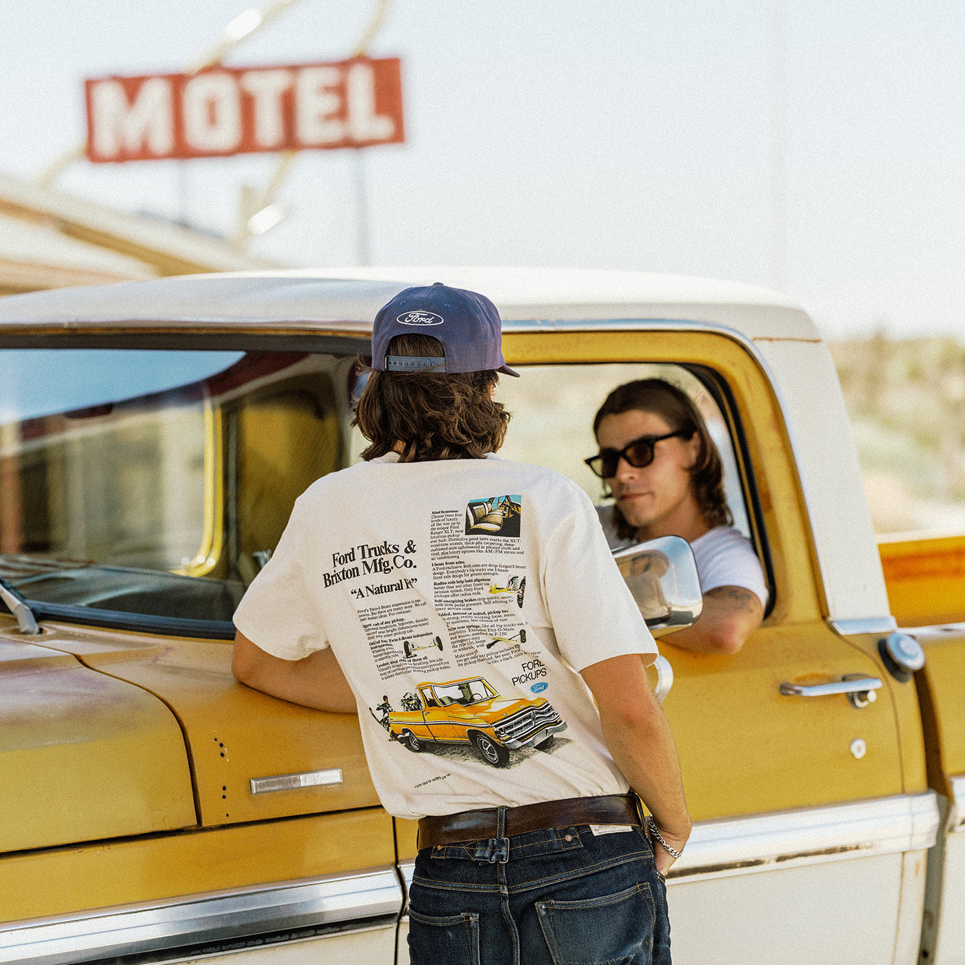 A person stands with their back to the camera, wearing a white graphic t-shirt with a vintage Ford Trucks print on the back, a blue Ford cap, and jeans, leaning on a yellow vintage truck next to a driver.