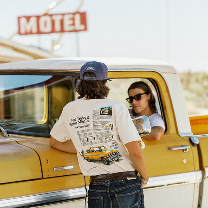 A person stands with their back to the camera, wearing a white graphic t-shirt with a vintage Ford Trucks print on the back, a blue Ford cap, and jeans, leaning on a yellow vintage truck next to a driver.