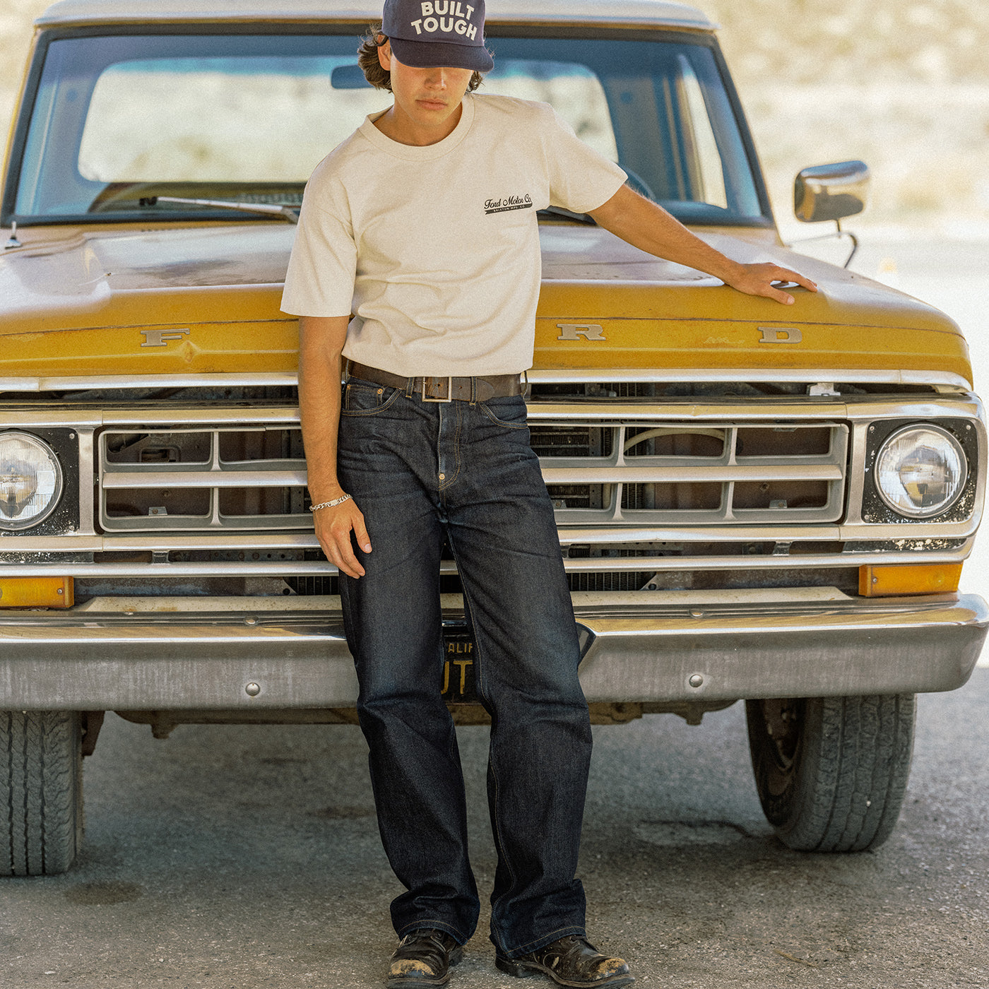 A man wearing a beige t-shirt with a 'Built Tough' logo, dark blue jeans, and a black cap stands in front of a vintage yellow pickup truck.