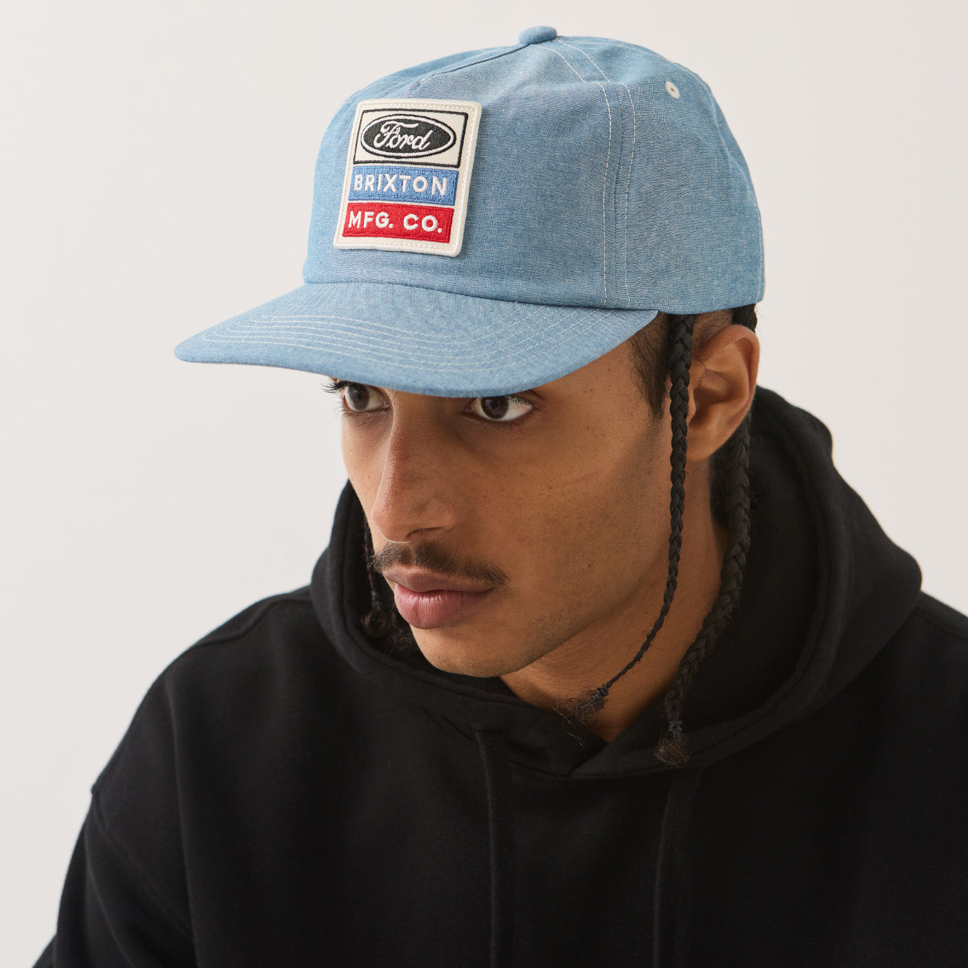 A man wearing a light blue Ford Brixton cap and a black hoodie, photographed against a plain light gray background.