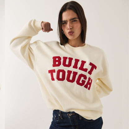 A woman modeling a cream-colored crewneck sweatshirt with 'BUILT TOUGH' printed in red across the chest, paired with dark blue jeans against a plain light background.
