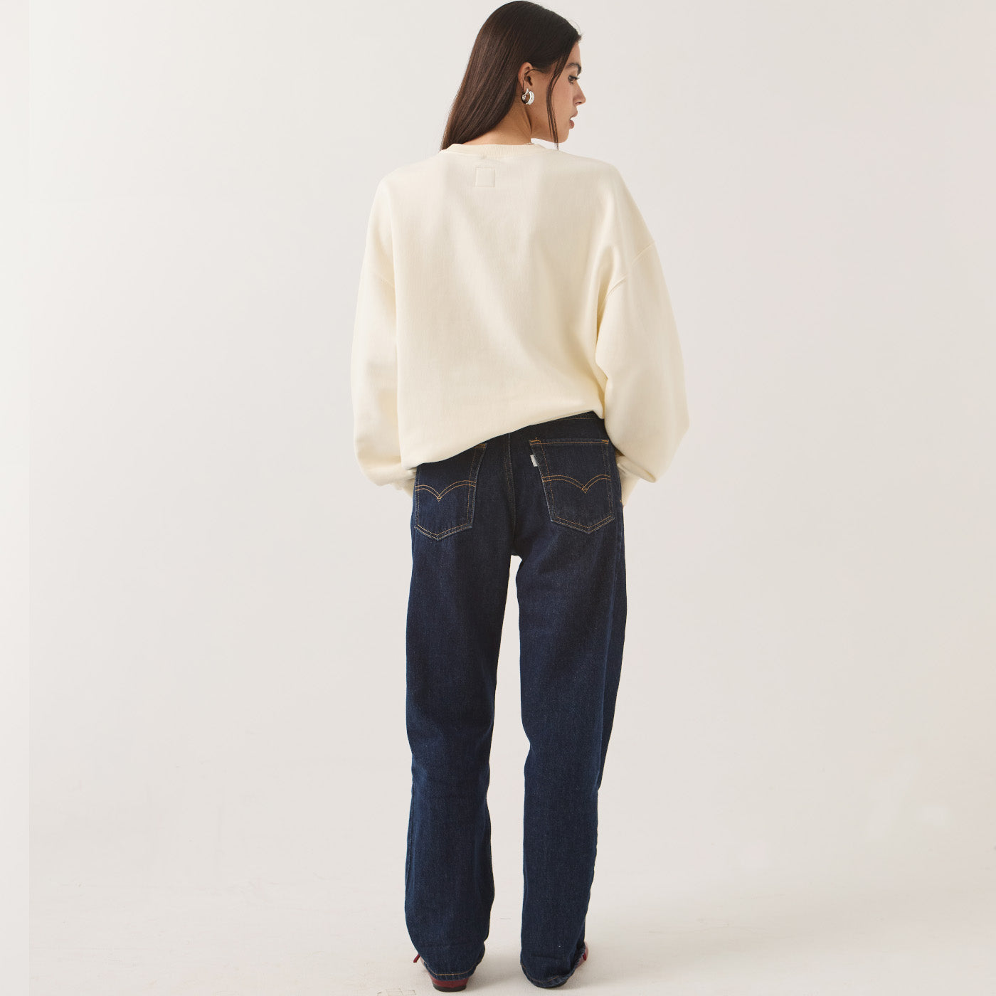 A woman shown from the back wearing a cream-colored, oversized long-sleeve knit sweater and dark blue straight-leg jeans against a plain off-white studio background.