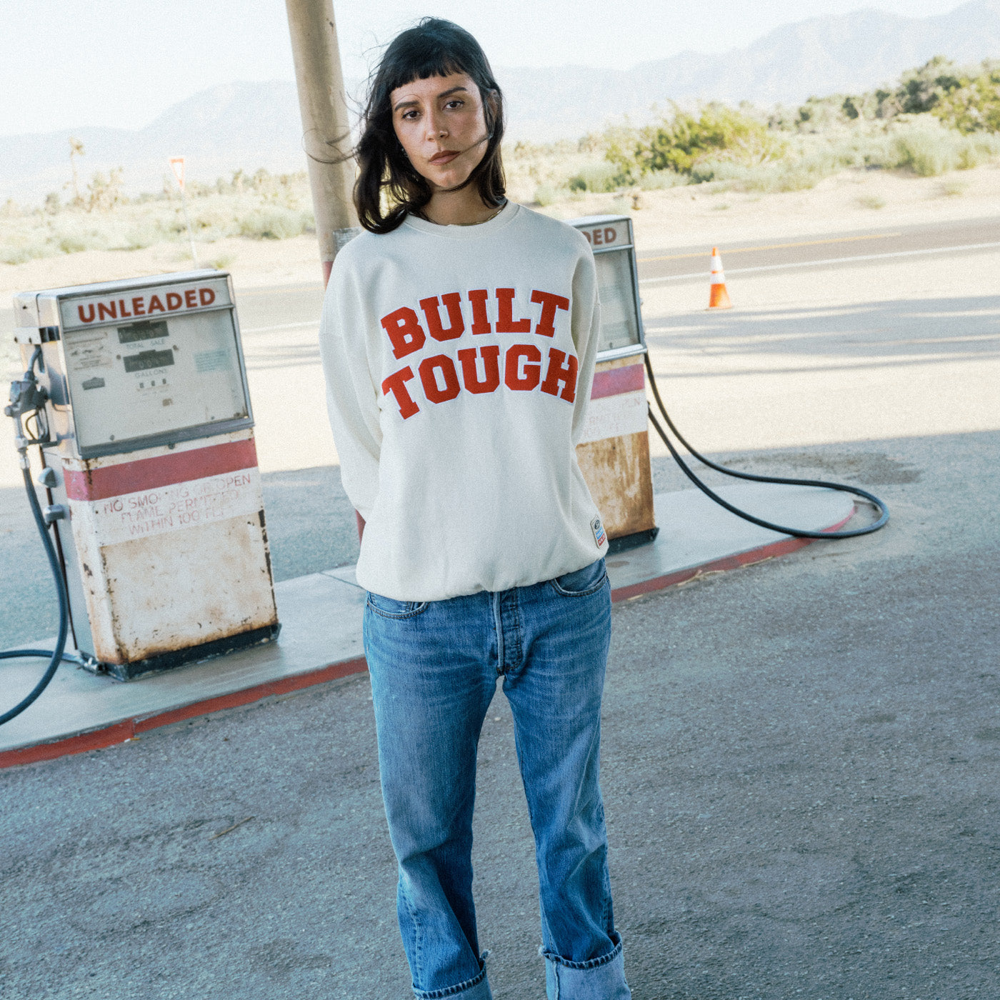A model stands at a vintage gas station wearing a white crewneck sweatshirt with 'BUILT TOUGH' printed in red across the chest, paired with blue jeans.