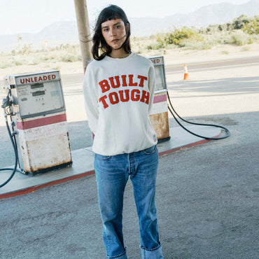 A model stands at a vintage gas station wearing a white crewneck sweatshirt with 'BUILT TOUGH' printed in red across the chest, paired with blue jeans.