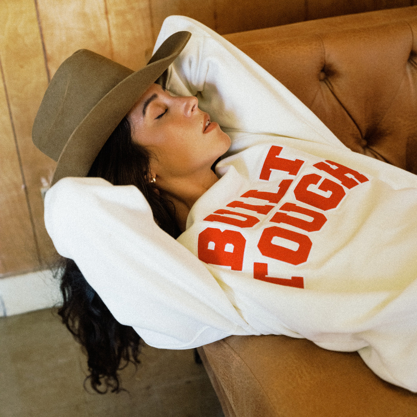 A woman wearing a brown wide-brim hat and a white sweatshirt with bold red text reading 'BUILT TOUGH' rests on a brown leather couch in a warm indoor setting.