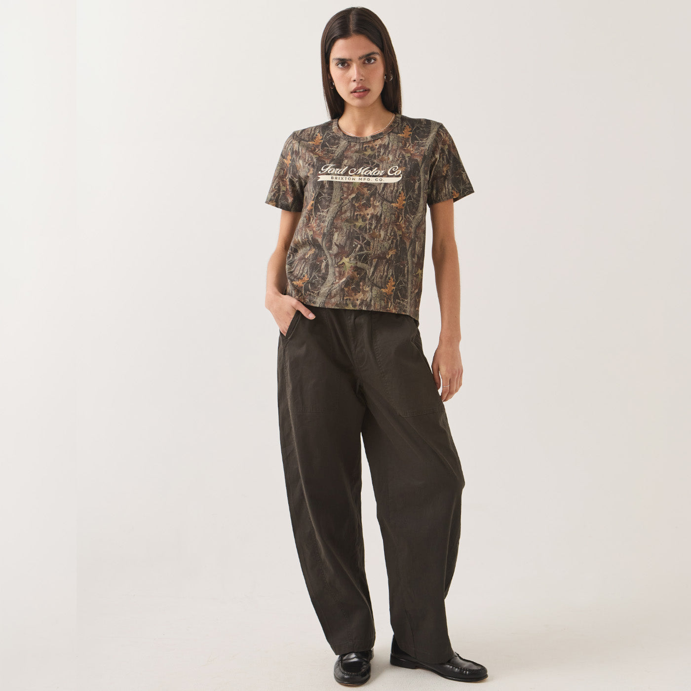 A model stands against a light gray studio background wearing a short-sleeve camouflage T-shirt with 'Realtree' printed across the chest and loose dark pants.