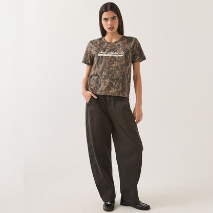 A model stands against a light gray studio background wearing a short-sleeve camouflage T-shirt with 'Realtree' printed across the chest and loose dark pants.
