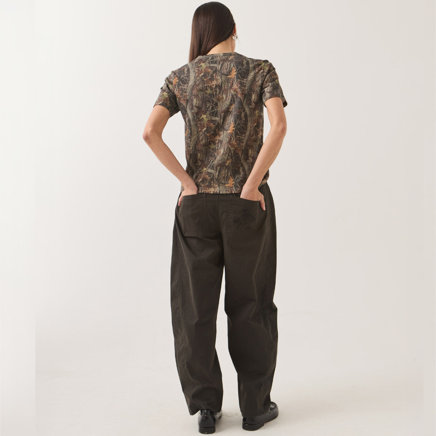A woman shown from the back wearing a short-sleeve camouflage-patterned top and dark wide-leg pants against a plain light background.