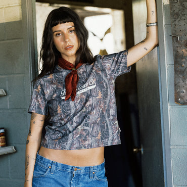 A woman with dark hair and bangs leans against a gray doorframe, wearing a short-sleeve cropped graphic t-shirt with a brown and gray camouflage pattern and a red neck scarf, paired with blue jeans.