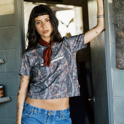 A woman with dark hair and bangs leans against a gray doorframe, wearing a short-sleeve cropped graphic t-shirt with a brown and gray camouflage pattern and a red neck scarf, paired with blue jeans.