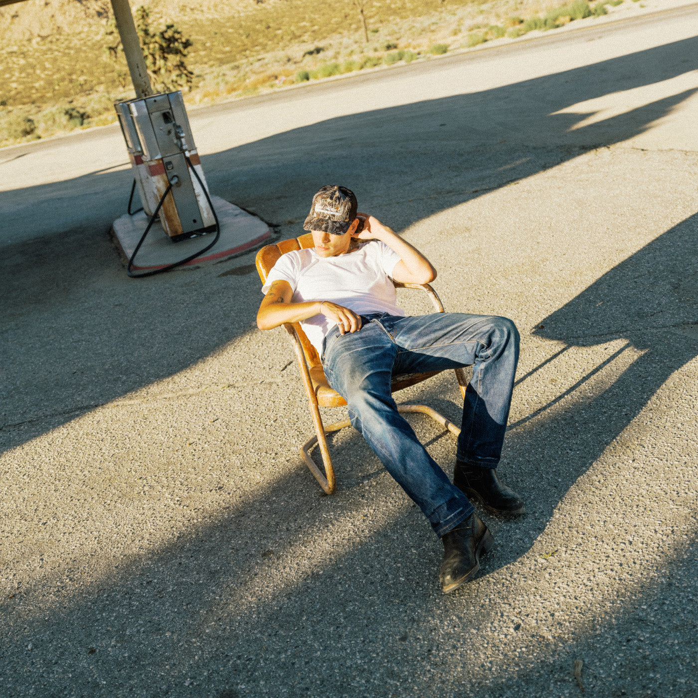 A man wearing a white t-shirt and blue jeans sits in a wooden folding chair on an asphalt road at a gas station, with a vintage gas pump visible in the background.
