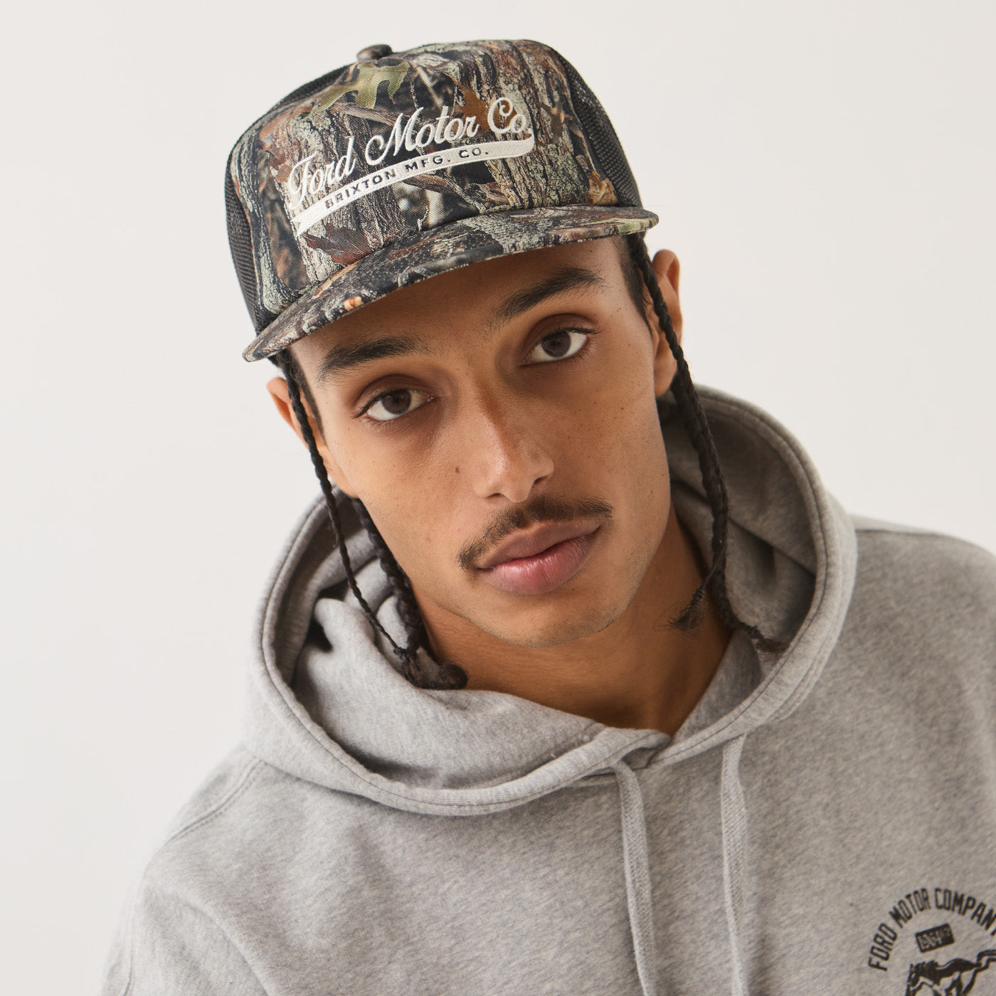 A man wearing a Ford Motor Company camouflage baseball cap and a gray hoodie with a visible Ford Motor Company logo on the chest, photographed against a plain light background.