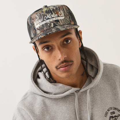A man wearing a Ford Motor Company camouflage baseball cap and a gray hoodie with a visible Ford Motor Company logo on the chest, photographed against a plain light background.