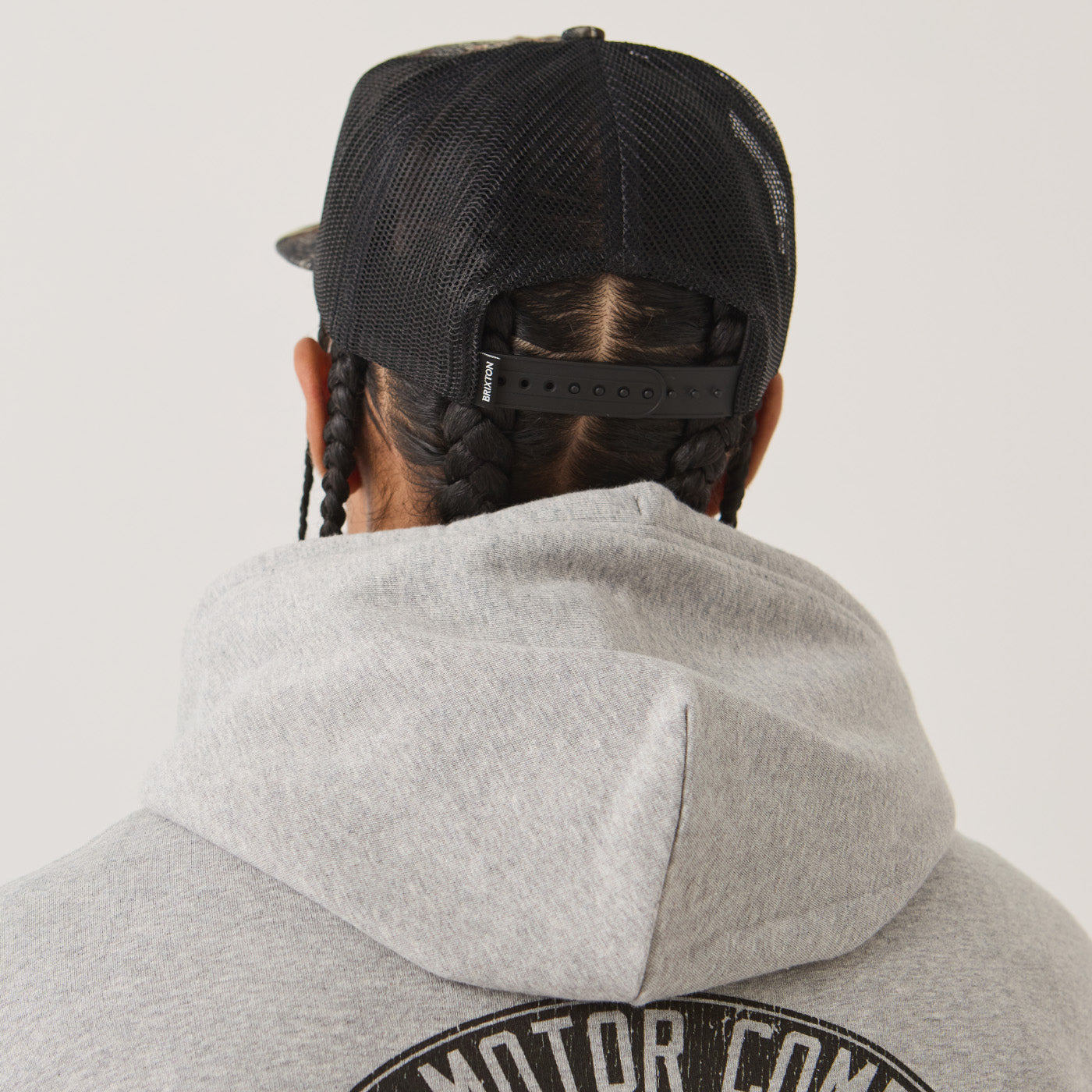 Rear view of a person wearing a black trucker cap and a light gray hoodie with a circular black graphic on the back.
