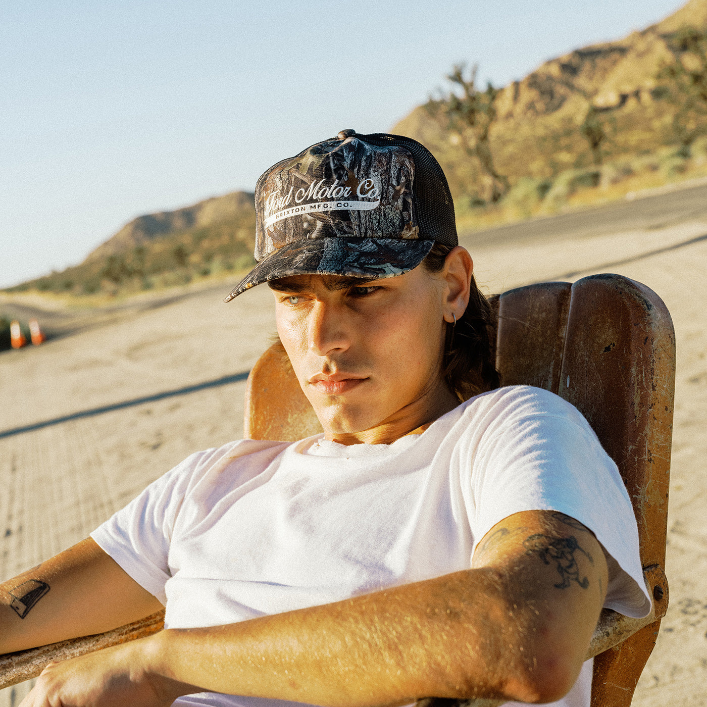 A man wearing a camouflage baseball cap with 'Real Motor Co' written on the front, sitting in a wooden chair outdoors on a sunny day.